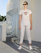 A woman wearing The Karlie white t-shirt with a heart and soul graphic by SUZI ROHER, along with white pants, sneakers, and sunglasses stands outdoors near a white architectural structure in bright sunlight.