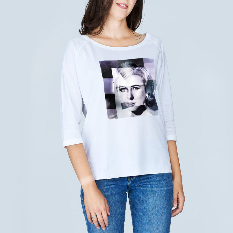 A woman models The Bailey tee by SUZI ROHER, featuring a boxy white cut, 3/4 sleeves, and a black-and-white graphic of a woman's face on the front. She's paired it with blue jeans and stands against a light gray background.
