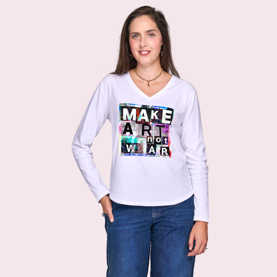 A creative woman with long brown hair, smiling at the camera, wears SUZI ROHER's The Victoria—a white long-sleeve shirt with bold "MAKE ART not WAR" lettering—paired with blue jeans against a pale pink background.