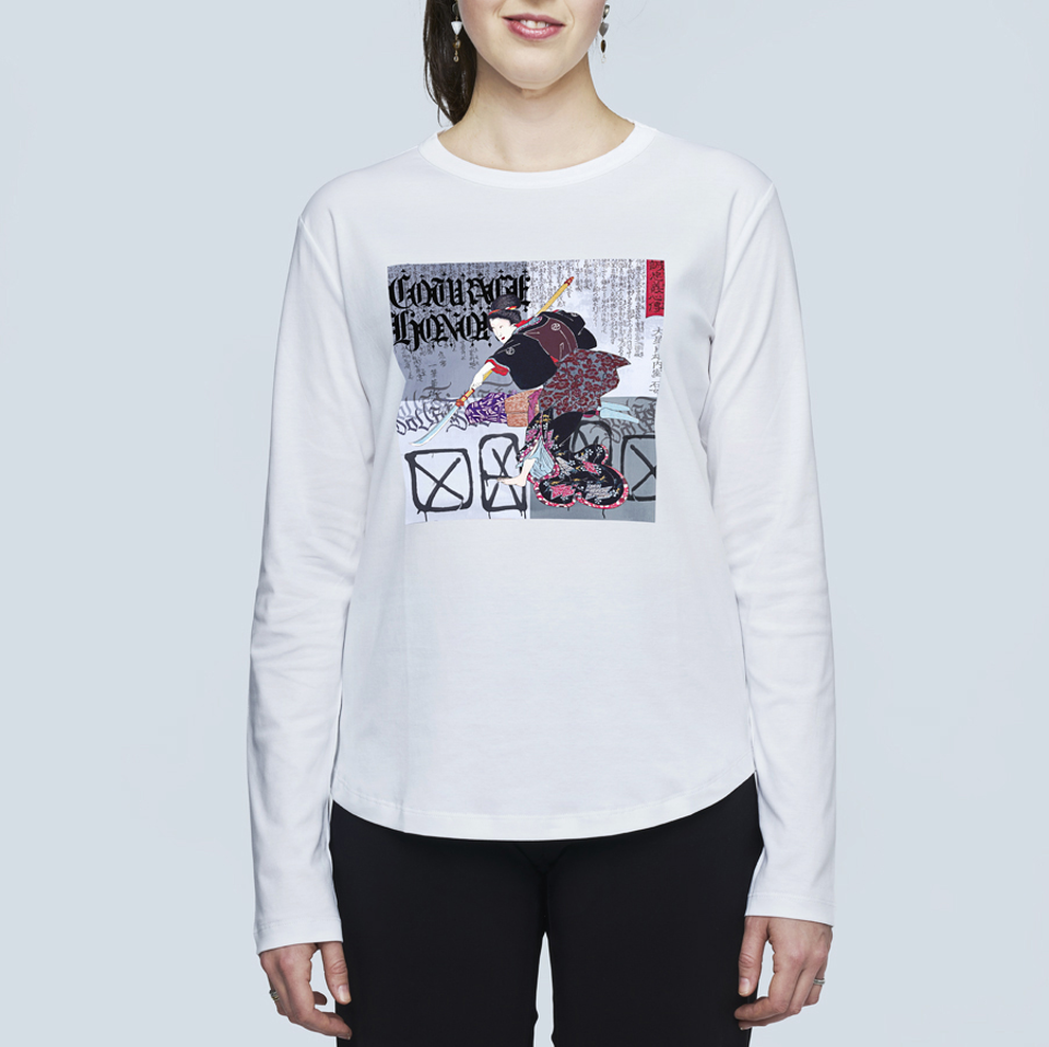 A person wears The Catherine by SUZI ROHER—a white long-sleeve shirt with an abstract female samurai graphic, bold text, and artistic elements—while standing against a light gray background.