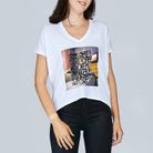 A woman wears SUZI ROHER’s The Velvet—a white oversized V-neck T-shirt with a colorful Coffee In Bed graphic and handwritten text—paired with black pants, standing against a light gray background.