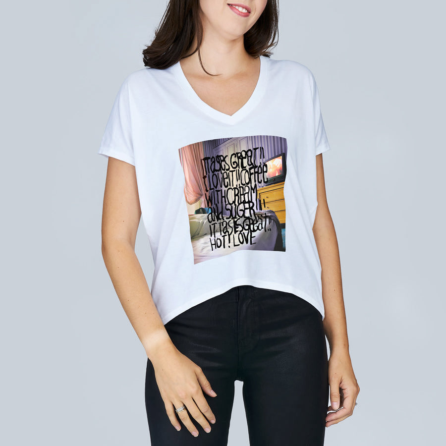 A woman wears SUZI ROHER’s The Velvet tee—a white, boxy-cut shirt with a vibrant Coffee In Bed graphic and black handwritten text: It feels great to love and be loved with charm and cheer. Styled here with black pants.