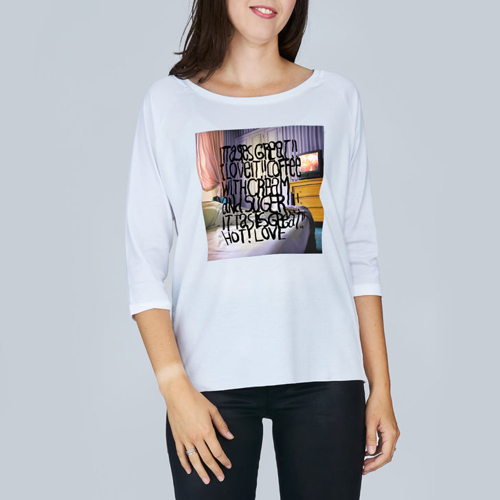 A woman smiles in front of a gray background, wearing The Bailey, a luxurious white long-sleeve shirt by SUZI ROHER featuring a graphic print and black text: "It’s great! I love the coffee with charm and sugar! It’s great! Not love!.