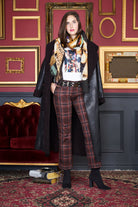 A woman wearing The Catherine by SUZI ROHER, a long black coat paired with plaid pants and heeled boots, stands confidently before an ornate red and gold Renaissance-style wall, next to a red velvet sofa and stacked books on the floor.