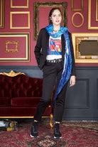 A woman poses confidently beside a red velvet sofa and gold frames, wearing The Bailey black suit by SUZI ROHER, a graphic Lucky Legs tee, blue patterned scarf, black sneakers, and jewelry in a chic red and gold room.