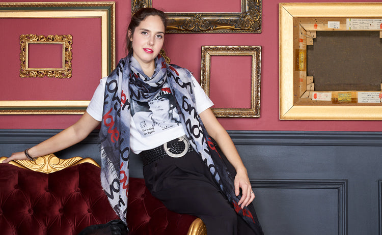 A stylish woman with dark hair in a ponytail sits on a red velvet sofa, wearing The Niki graphic t-shirt by SUZI ROHER, black pants, and a patterned scarf. Ornate empty picture frames decorate the red and gray wall behind her.