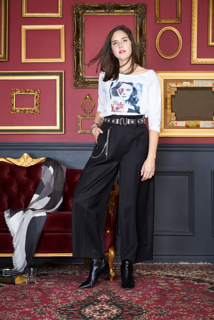 A woman poses indoors by a red velvet couch, wearing The Bailey wide-leg pants from SUZI ROHER with a graphic tee and black boots. She stands on a patterned rug against red walls with empty gold picture frames.