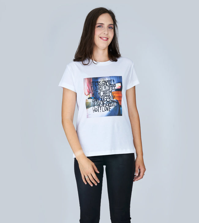 A woman with long brown hair, wearing The Niki graphic tee by SUZI ROHER featuring "Coffee In Bed" text and a colorful design, pairs it with black pants and smiles against a plain light gray background.