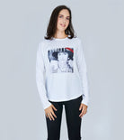 A woman with long brown hair wears SUZI ROHER’s The Catherine, a slim fit white long-sleeve shirt featuring a black-and-white graphic of a woman in a hat and jewelry, standing against a light gray background with her hands by her sides.