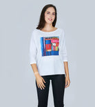 A woman with long brown hair smiles softly, wearing SUZI ROHER's The Bailey—a white boxy cut tee featuring a colorful book spines graphic—paired with black pants, standing against a light gray background.