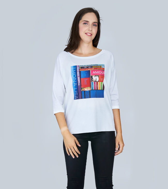 A woman with long brown hair smiles softly, wearing SUZI ROHER's The Bailey—a white boxy cut tee featuring a colorful book spines graphic—paired with black pants, standing against a light gray background.
