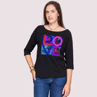 A woman with long brown hair wears SUZI ROHER’s The Bailey, a black boxy cut tee featuring a colorful geometric LOVE graphic, paired with blue jeans and standing against a plain light background.