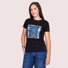 A woman with long brown hair, wearing The Niki black 100% cotton graphic t-shirt by SUZI ROHER and blue jeans, stands against a light pink background, gazing at the camera with a relaxed expression.