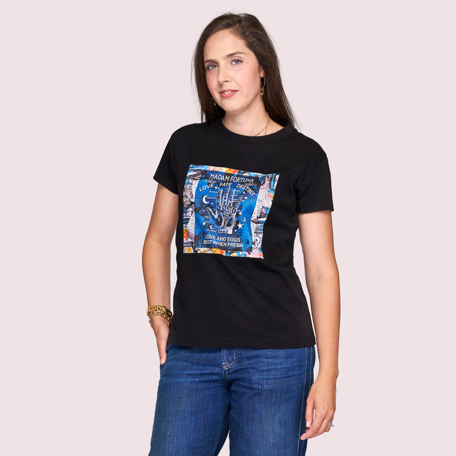 A woman with long brown hair, wearing The Niki black 100% cotton graphic t-shirt by SUZI ROHER and blue jeans, stands against a light pink background, gazing at the camera with a relaxed expression.
