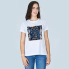 A woman with straight brown hair wears SUZI ROHER’s The Niki tee—100% cotton, crew neck, white with a colorful "Madam Fortuna Love Fate Destiny" graphic—paired with blue jeans, standing against a light gray background.