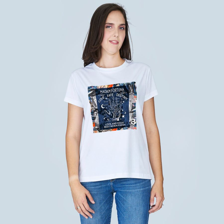 A woman with straight brown hair wears SUZI ROHER’s The Niki tee—100% cotton, crew neck, white with a colorful "Madam Fortuna Love Fate Destiny" graphic—paired with blue jeans, standing against a light gray background.