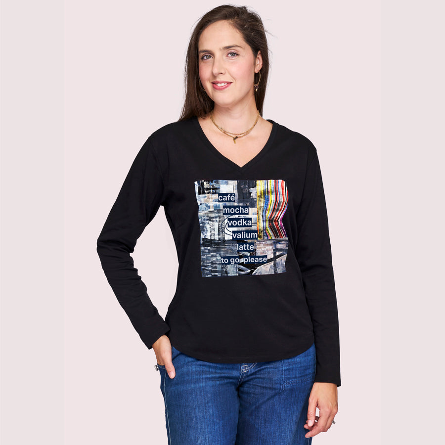 A woman with long brown hair smiles with one hand in her jeans pocket, wearing SUZI ROHER's "The Victoria"—a black long-sleeve V-neck shirt featuring a colorful graphic and supportive words, against a plain light background.