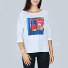 A woman wears The Bailey tee by SUZI ROHER, featuring a white boxy cut and a colorful book spine print with titles like “MONA LISA” and “AMOUR,” posed against a plain gray background.