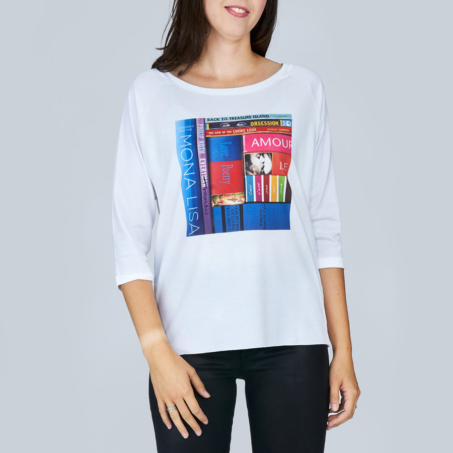 A woman wears The Bailey tee by SUZI ROHER, featuring a white boxy cut and a colorful book spine print with titles like “MONA LISA” and “AMOUR,” posed against a plain gray background.