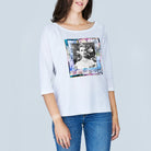 A woman wears The Bailey by SUZI ROHER—a white, 100% cotton, boxy tee with a colorful square graphic of vintage Audrey Hepburn and text—paired with blue jeans against a light gray background.