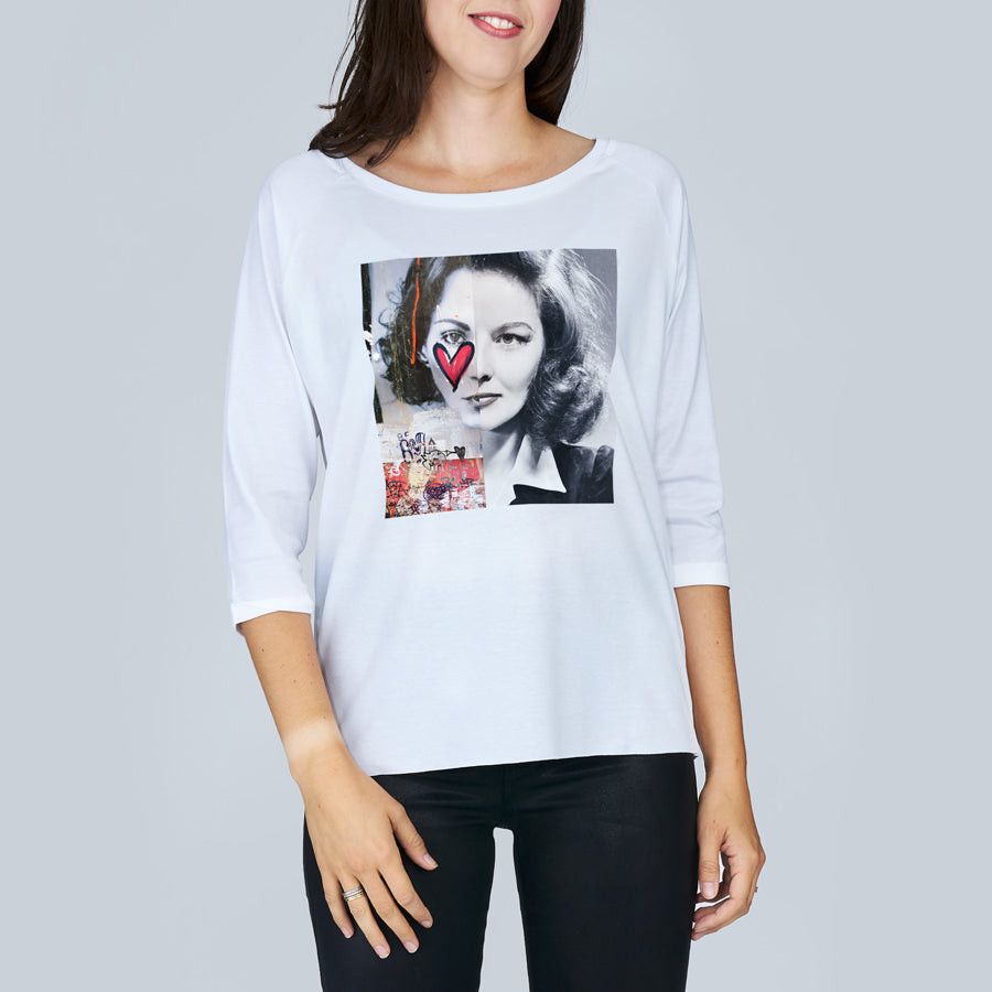 Smiling, a woman wears The Bailey by SUZI ROHER—a white boxy tee featuring a black-and-white portrait with red heart and collage accents, echoing timeless Hepburn fashion.