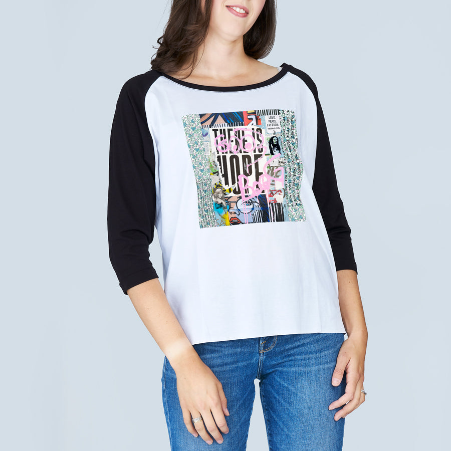 A woman stands against a light gray background wearing SUZI ROHER's The Bailey, a 100% cotton boxy cut tee featuring a colorful abstract graphic and "There is Hope" text on the front, paired with blue jeans.