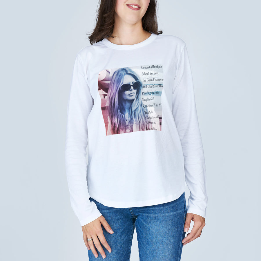 A woman stands smiling in The Catherine by SUZI ROHER—a slim fit, 100% cotton white long-sleeve tee with a graphic of a woman in sunglasses and multi-font text on the front, paired with blue jeans against a light gray background.