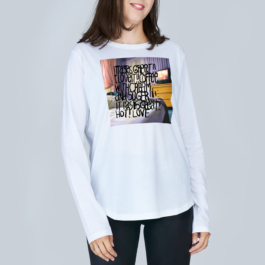 A woman smiles in SUZI ROHER’s The Catherine, a long-sleeved 100% cotton crew neck tee featuring a colorful Coffee In Bed graphic and handwritten text, standing against a plain light gray background. Her face is partially visible.