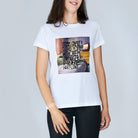A woman stands against a plain light background wearing The Niki by SUZI ROHER, a white t-shirt featuring a colorful graphic and handwritten-style text that celebrates the luxurious joy of Coffee In Bed with cream and sugar.