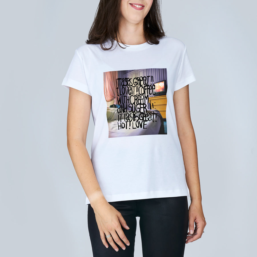 A woman stands against a plain light background wearing The Niki by SUZI ROHER, a white t-shirt featuring a colorful graphic and handwritten-style text that celebrates the luxurious joy of Coffee In Bed with cream and sugar.