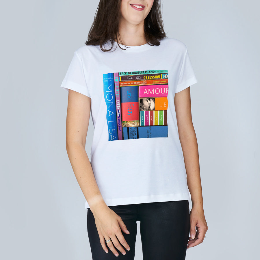A woman is shown from the torso up, smiling and wearing SUZI ROHER's The Niki t-shirt, which features colorful, upright book spines on the front. The background is plain light gray.