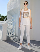 Wearing SUZI ROHER’s The Lola—sleeveless white graphic top—a woman in sunglasses, white pants, and sneakers stands outdoors by a sunlit white building with a striking circular feature, ready for adventure.