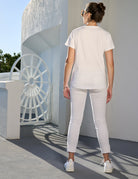 A woman stands outdoors, facing away, embodying iconic grace in The Viv white t-shirt by SUZI ROHER. Near a circular gate and curved wall, she channels Jackie Kennedy as sunlight casts gentle shadows on the ground.