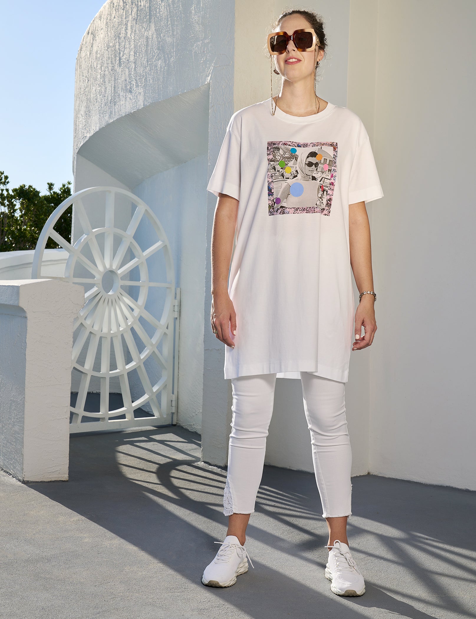 A woman wearing sunglasses and the SUZI ROHER "Daisy, Because We Love You" oversized essential long white t-shirt with a colorful graphic stands outdoors in bright sunlight near a white building, paired with white pants and sneakers.