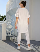 A woman in a SUZI ROHER "Daisy, Because We Love You" white oversized tunic stands outdoors, back to the camera, facing a round white structure. She pairs it with sunglasses, cropped pants, and sneakers in the bright sunlight.