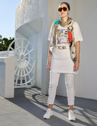 Wearing SUZI ROHER's "Daisy, Because We love You" tunic dress, a woman in sunglasses, white pants, sneakers, and a patterned scarf stands confidently by a decorative white wall and sunlit wheel-shaped window.
