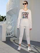A creative woman in sunglasses, white pants, sneakers, and SUZI ROHER’s The Victoria long-sleeve shirt stands outside by a striking white structure with a large wheel design—an inspiring scene.