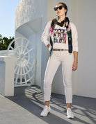 A stylish woman in sunglasses stands outside, wearing The Victoria scarf by SUZI ROHER with a white tee, jeans, and sneakers. She radiates inspiration in front of a modern white building with a circular design.