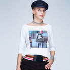 A woman in a navy cap, silver choker, and bracelet poses in SUZI ROHER's The Bailey boxy cut tee with a Bette Davis graphic, black pants, and striped belt. Red lipstick and striking blue eyes complete her look.