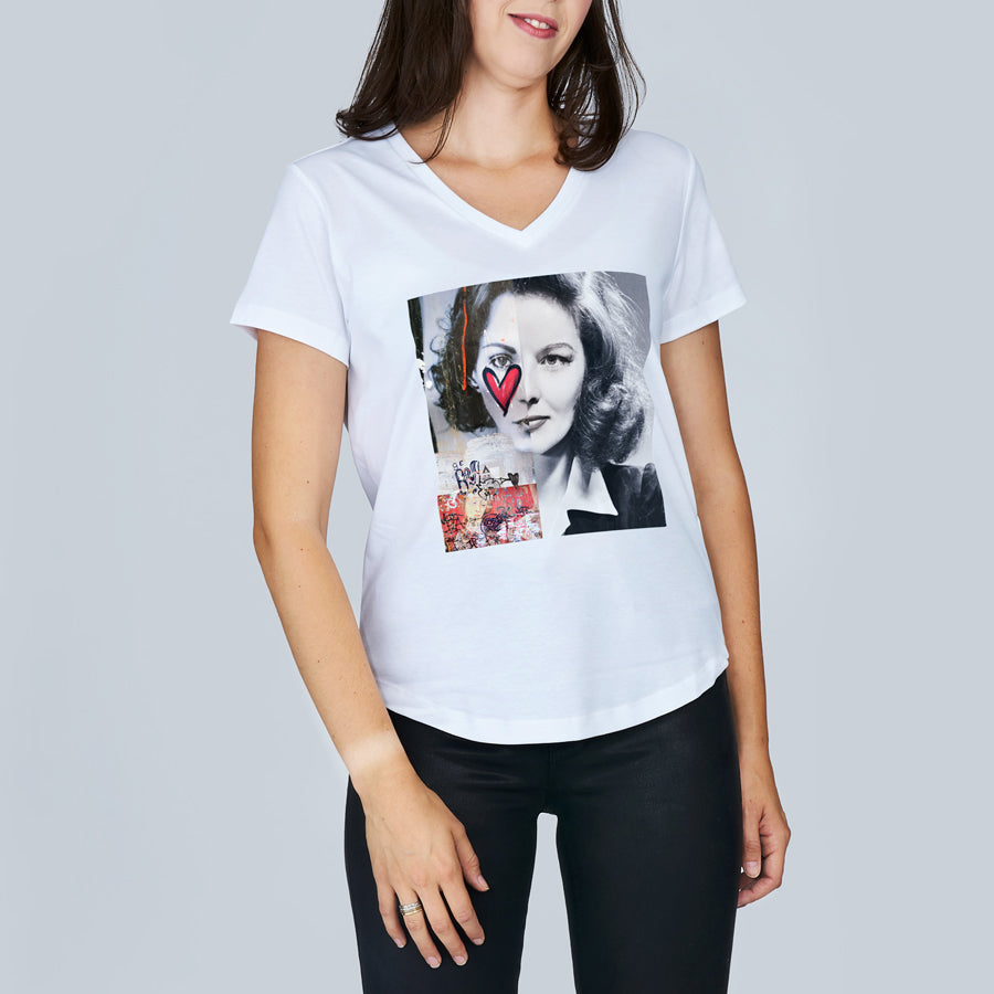 A woman models SUZI ROHER's The Viv— a slim fit, 100% cotton V-neck tee with an artistic black-and-white collage, colorful accents, and a red heart on the front—paired with black pants against a plain background.