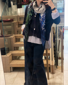 A person snaps a mirror selfie in a store, styled in SUZI ROHER’s A La Mode silk scarf—featuring green, blue, and gray patterns—paired with wide-leg dark pants, a dark blazer, and striped shirt. Their face is hidden by the phone.
