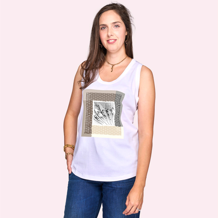 A woman with long brown hair smiles against a light pink background, wearing The Lola white sleeveless graphic top by SUZI ROHER and blue jeans, as if she's just enjoyed her first surf lesson.