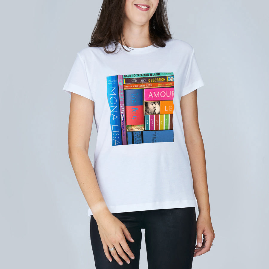 A woman stands against a plain light background, wearing The Niki by SUZI ROHER—a white t-shirt with a colorful book spine print featuring titles like Mona Lisa, Amour, and Obsession, celebrating the mysteries of life.