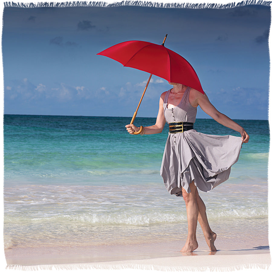 Wearing SUZI ROHER's "Barefoot On The Beach," a person in a gray dress and scarf stands barefoot on the sand, holding a red umbrella as the turquoise ocean shimmers under a partly cloudy sky, their face turned away.