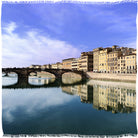 A row of colorful buildings lines the riverbank, their reflections shimmering as an arched stone bridge spans the water beneath a partly cloudy sky—the ideal scene to enjoy SUZI ROHER's Campari Time.